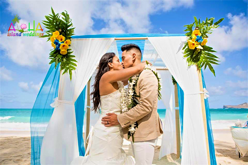Couple kissing under a blue and white beach arch decorated with yellow flowers